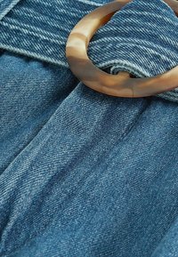 Denim fabric with a blue hue featuring a wide waistband and a large, translucent tortoiseshell-style circular buckle accent.