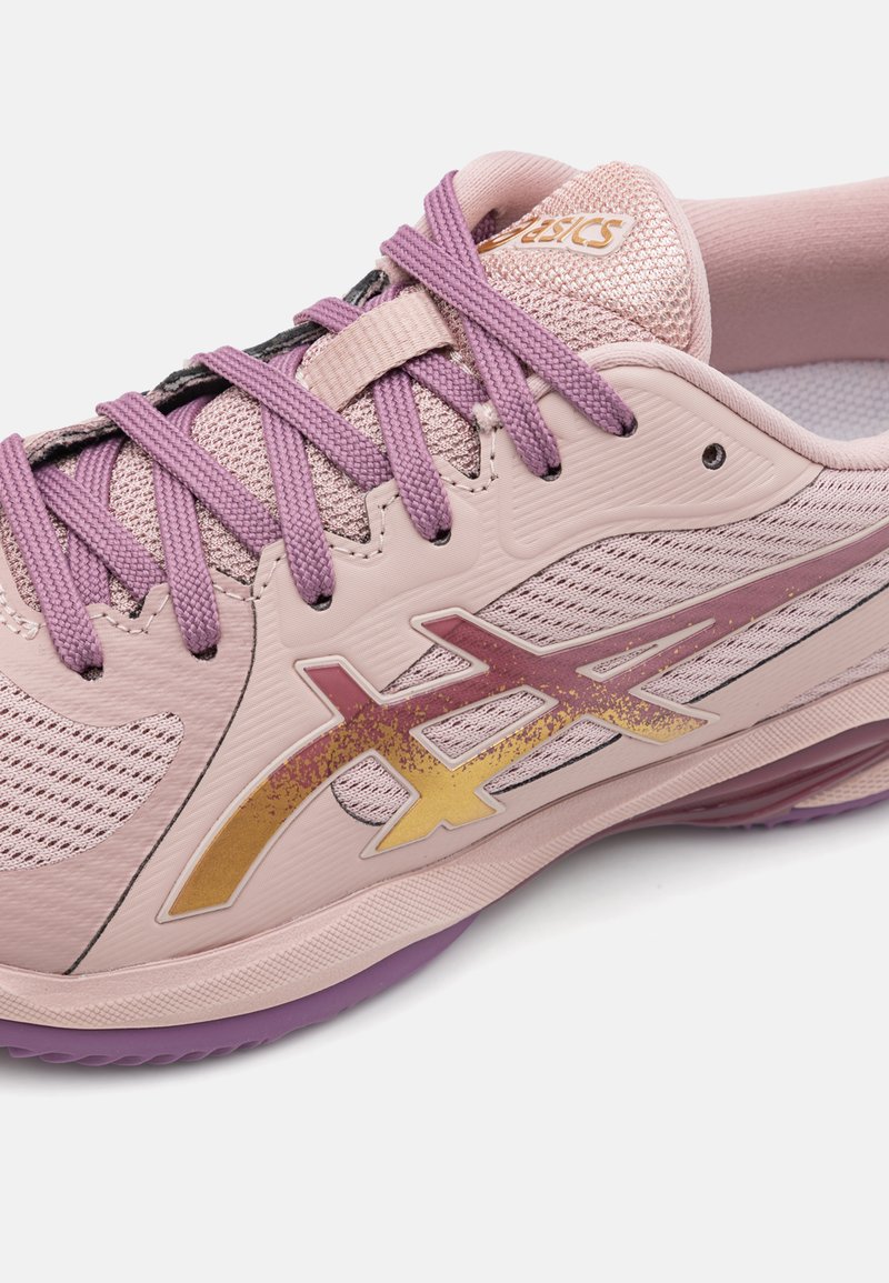 Zapatilla deportiva de color rosa claro con acentos morados, parte superior de malla transpirable, diseño decorativo en capas y detalle del logo dorado en el lateral.