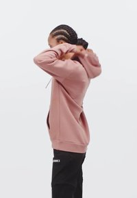 Felpa rosa chiaro con cappuccio e cordino, tasche laterali e polsini a coste; indossata con pantaloni neri con un logo sulla gamba.