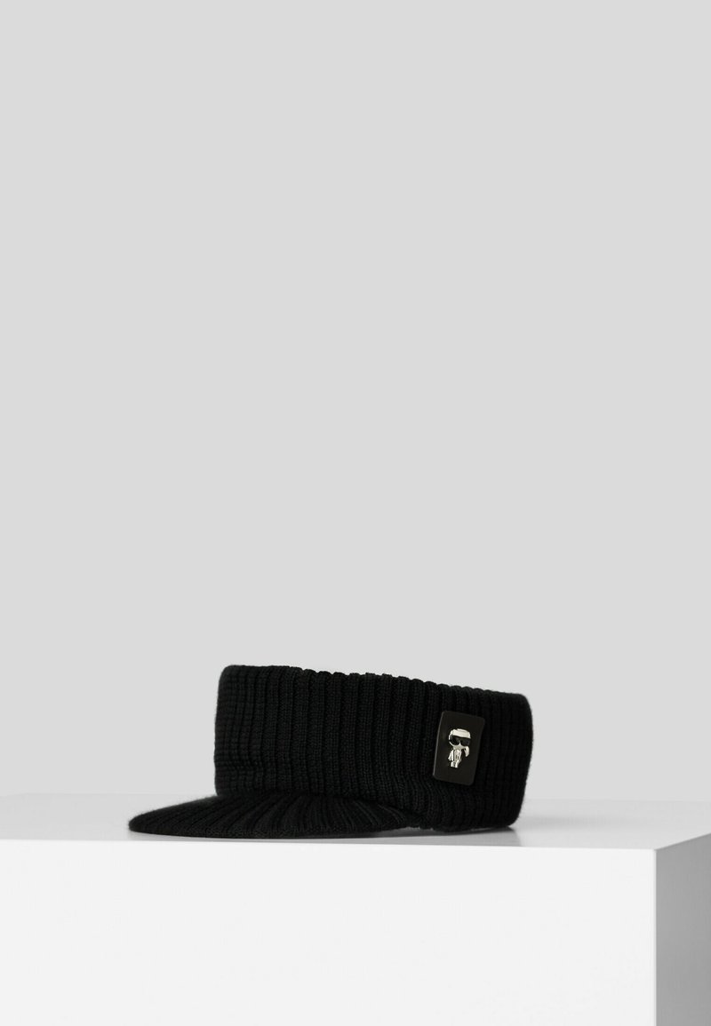 Casquette à visière noire en tricot côtelé avec un petit emblème argenté représentant une figure stylisée sur le côté.