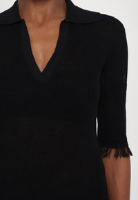 Haut en maille noire avec un décolleté en V et des manches courtes. Présente un col et des détails à franges aux extrémités des manches. Texture légère et semi-transparente.