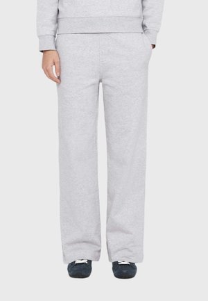 Person wearing light gray sweatpants and dark sneakers with white laces, standing with one hand in pocket against a plain background.