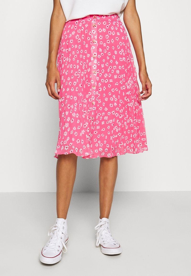 Jupe rose, mi-longue, avec un motif floral, dotée de boutons sur le devant et en tissu léger et fluide. Portée avec des baskets blanches.