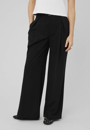 Femme portant un pantalon large taille haute noir avec plis, mains dans les poches, assorti de ballerines noires cloutées.