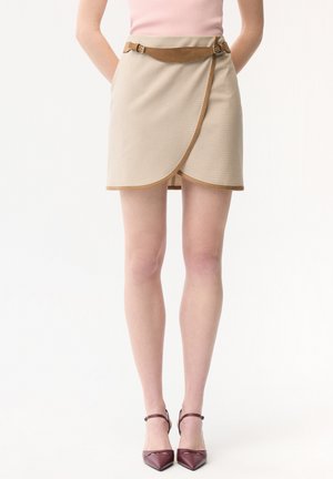 Femme portant une jupe portefeuille beige avec une ceinture marron et des escarpins marron à bouts pointus, debout les mains derrière le dos sur un fond blanc.