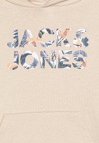 Béžová mikina s kapucňou a predným vreckom, na ktorom je nápis "JACK & JONES" v vyrezávaných písmenách vyplnených kvetinovým motívom v modrej, zelenej a korálovej farbe.