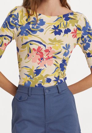 Femme portant un haut floral ajusté avec des fleurs bleues, roses et jaunes, associé à un pantalon bleu taille haute avec des poches boutonnées.