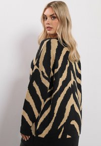 Blond kvinna som ser över axeln, iklädd en svart och beige zebra-randig tröja mot en ljus, enfärgad bakgrund.
