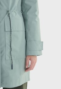 Veste imperméable bleu clair avec une taille à cordon, deux poches avant et un poignet carré. Texture lisse avec des détails de couture visibles.