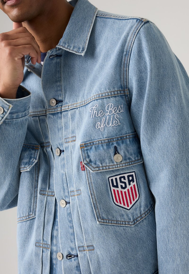Homme portant une veste en jean bleu clair avec un texte brodé "The Best of U.S." et un écusson en forme de bouclier des États-Unis sur la poche avant.