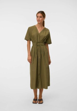 Femme portant une robe midi vert olive à manches courtes, col en V et taille nouée, associée à des sandales ouvertes foncées, debout sur un fond uni.