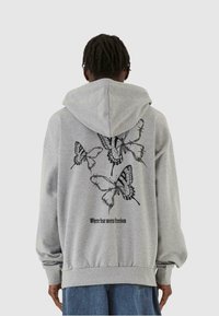 Felpa grigia con cappuccio, presenta grafiche di farfalle nere e la scritta "Dove la paura incontra la libertà" sul retro, polsini e orlo a costine.