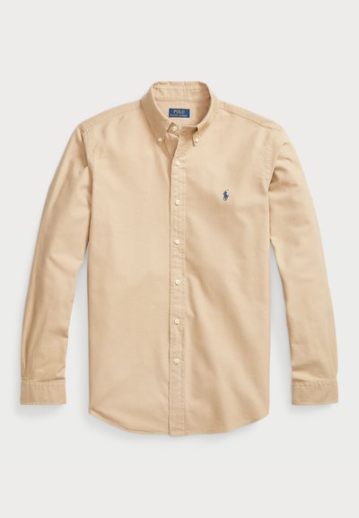 Hele beež puhvärava särk puuvillast, klassikalise kraega, pikkade varrukatega, valgete nööpidega ja väikese sinise logoga rinnal.