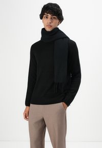 Man in een zwarte trui en sjaal met beige broek, staande met één hand in de zak tegen een effen witte achtergrond.