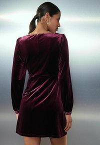 Robe en velours d'un bourgogne profond, à longues manches avec des épaules froncées, taille cintrée, jupe évasée, et poignets accentués avec des détails.