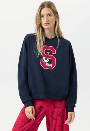 Vrouw in een marineblauwe sweatshirt met een rode "S" en een grafiek van Snoopy en Woodstock, gecombineerd met rode broek met zwarte Peanuts-karakterprints.