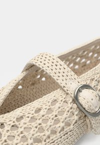 Chaussure en tissu beige tissé avec un design ouvert, dotée d'une bride décorative et d'une boucle argentée. Surface texturée avec un motif en maille.