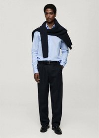 Cămașă button-up de un albastru deschis, pulover tricotat navy așezat pe umeri, pantaloni negri și pantofi negri. Design simplu cu texturi variate.