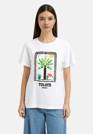 Junge Frau trägt ein weißes T-Shirt mit einem bunten Palmenmotiv und dem Text „Tulum Mexico“, kombiniert mit hellblauen Jeans.