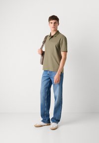 Olivgrünes Poloshirt mit Kragen, kombiniert mit hellblauen geraden Jeans und weißen Sneakers mit braunen Akzenten. Hält eine graue Messenger-Tasche.