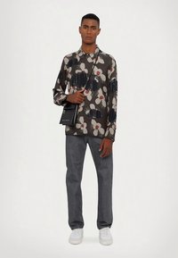 Moški v srajci s cvetličnim vzorcem, sivi kavbojki, belih supergah in z črno torbo čez ramo, v nasprotju z enostavno ozadjem.