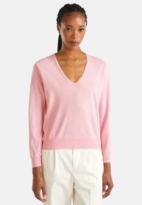 Mujer con trenzas que lleva un suéter de pico en rosa claro y pantalones blanco roto, de pie contra un fondo claro y liso.