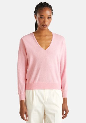 Vrouw met vlechten in een lichtroze V-hals trui en off-white broek, staand tegen een effen lichte achtergrond.