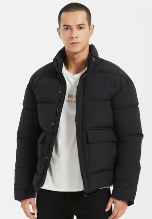 Doudoune noire avec fermeture éclair, col à boutons pression, poches latérales et texture matelassée. Portée par-dessus une chemise blanche. Coupe décontractée.