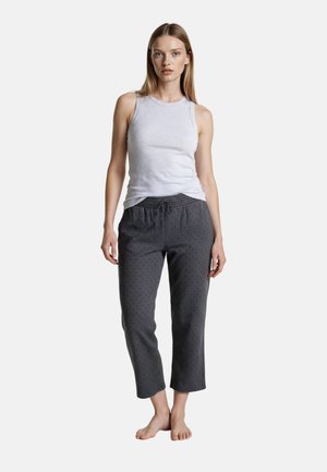 Žena stojící naboso, oblečená v bílém tílku a tmavě šedých zkrácených kalhotách s jemným vzorem na jednobarevném pozadí.