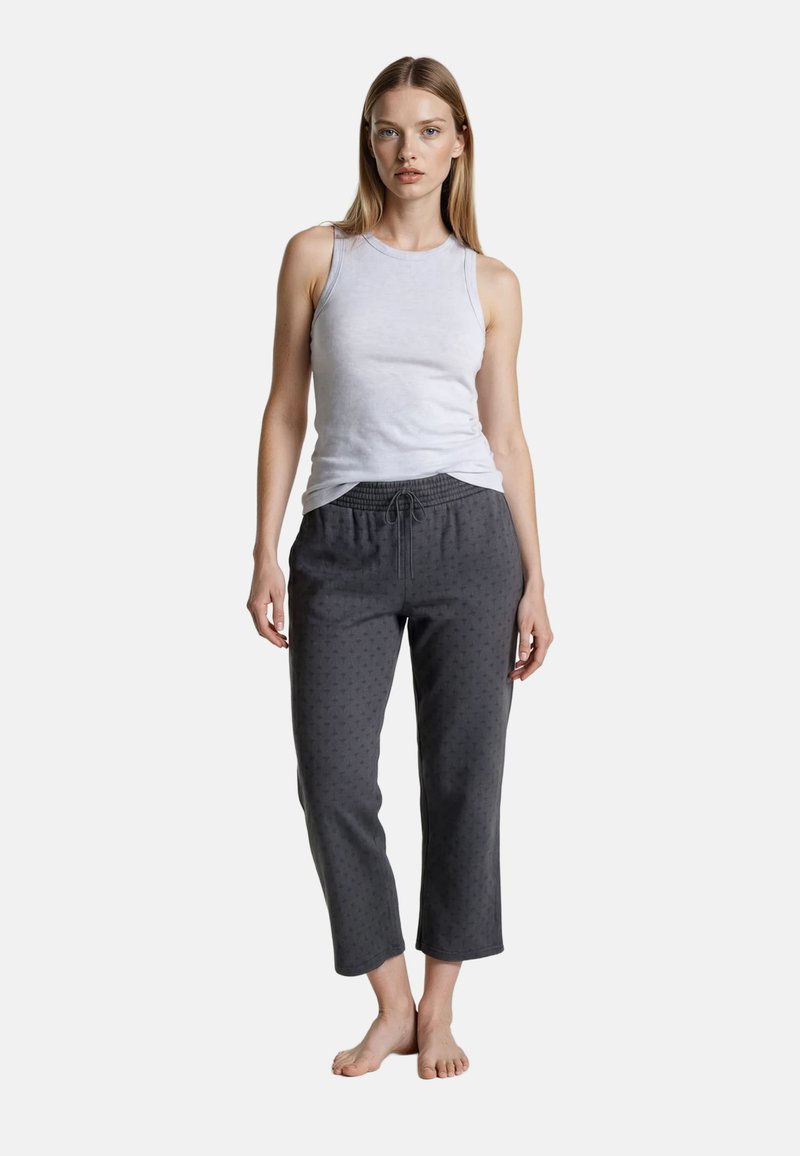 Kvinde stående barfodet iført en hvid ærmeløs top og mørkegrå kapribukser med et diskret mønster på en ensfarvet baggrund.