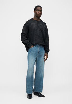 Uomo in piedi con la mano sinistra nella tasca dei jeans blu a gamba larga, indossa una camicia nera a maniche lunghe con motivo e stivali neri a punta su sfondo bianco.