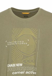 Oliwkowy bawełniany T-shirt z okrągłym dekoltem, żółtym konturowym grafiką oraz białym tekstem: "NIESKOŃCZONY POSZUKIWACZ PRZYGÓD" i "GONI NOWE HORYZONTY."