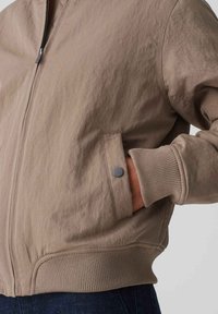 Giacca bomber marrone chiaro con finitura testurizzata, polsini a costine e tasca con bottoni a pressione sul lato sinistro. Chiusura frontale con zip.