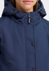 Veste bleu marine avec fermeture éclair et pression, dotée d'une capuche réglable et d'une texture côtelée sur le tissu.