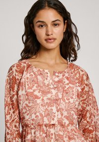 Blus med blommönster i ljus persika med intrikata orangea mönster. Har en rundad halsringning och knappstängning, tillverkad av lättviktig tyg.