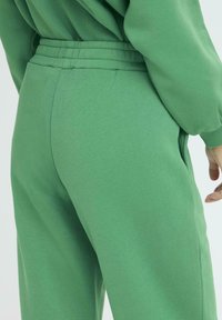 Zelene pamučne trenirke s elastičnom trakom i bočnim džepovima. Tkanina izgleda mekano i udobno, s glatkom teksturom.