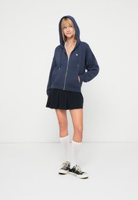 Marineblauwe ritssluiting hoodie met een gevoerde capuchon, voorkruiszakken en een klein logo; gecombineerd met een zwarte geplooide rok en witte kniemediumsokken.