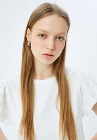 Witte mouwloze top met een gladde textuur en geborduurde oogjesdetails op de mouwen, gecombineerd met lang steil haar en studs.
