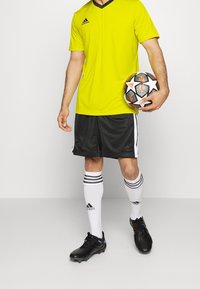 Jersey amarillo de manga corta con acentos negros, pantalones cortos de malla negros, calcetines blancos con rayas negras y botas de fútbol negras, sosteniendo un balón de fútbol con un diseño.