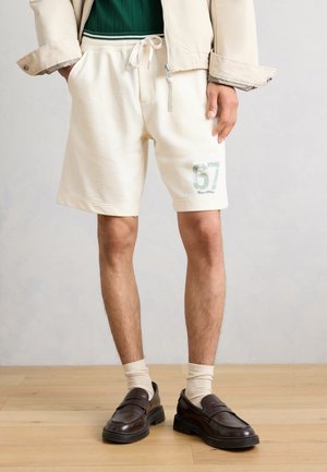 Személy, aki krémszínű, húzózsinóros rövidnadrágot visel „67” és a Marc O'Polo logóval, bézs dzsekit, krémszínű zoknit és sötétbarna loafereket fa padlón.