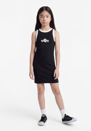 Νεαρή κοπέλα που στέκεται, φορώντας αμάνικο μαύρο φόρεμα με λευκό σχέδιο λουλουδιών, λευκές κάλτσες και μαύρα sneakers χαμηλού ύψους σε λευκό φόντο.