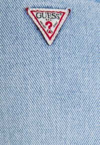Tissu en denim bleu clair avec un patch logo GUESS visible en forme de triangle, présentant des accents rouges et blancs. Surface texturée.