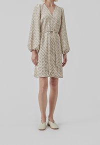 Hellbeiges Kleid mit floralem Druck, V-Ausschnitt, elastischen Ballonärmeln, geraffter Taille und knielanger Länge. Kombiniert mit hellgrünen Slippern.