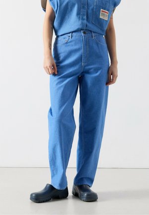 Osoba oblečená v svetlomodrých džínsových nohaviciach s vysokým pásom, zodpovedajúcej bezrukávovej džínsovej košeli a čiernych nazúvacích topánkach, stojaca na svetlej podlahe.