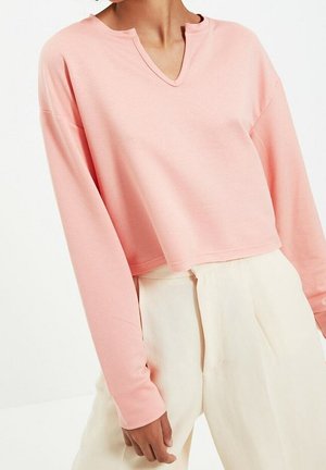 Kvinde iført en lyserød cropped top med lange ærmer med en hakket halsudskæring og højtaljede, off-white plisserede bukser.