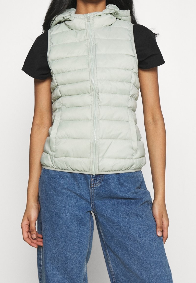 Femme portant une veste matelassée sans manches à capuche gris clair sur un t-shirt noir à manches courtes et un jean bleu taille haute, debout devant un fond blanc.