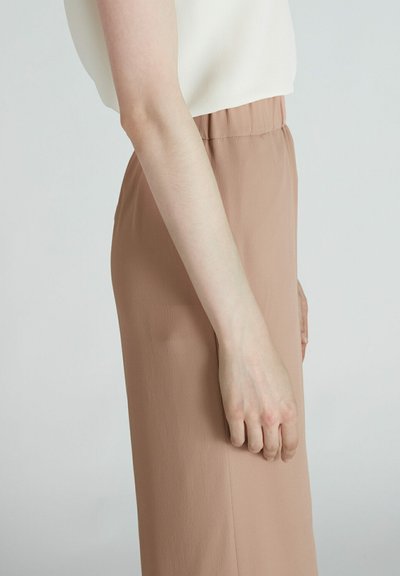 Licht beige elastische taillebandbroeken met een gladde textuur, vanuit de zijaanzicht weergegeven, gecombineerd met een wit topje. Er zijn geen zichtbare patronen.