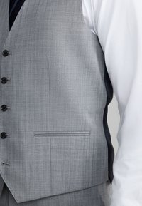 Közelkép egy személyről, aki világosszürke gombos mellényt visel fehér hosszú ujjú ing felett, bemutatva a szövet textúráját és a mellény elejének részleteit.