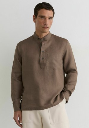 Bărbat purtând o cămașă maro cu mâneci lungi, guler mandarin și nasturi, asortată cu pantaloni deschiși la culoare, stând în fața unui perete simplu.