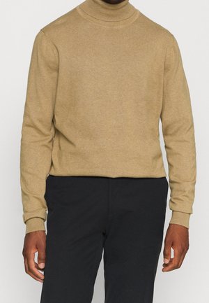 Homme portant un pull à col roulé beige et un pantalon noir, debout les mains détendues le long du corps devant un fond uni.